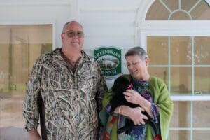 An old couple standing and holding a small black puppy